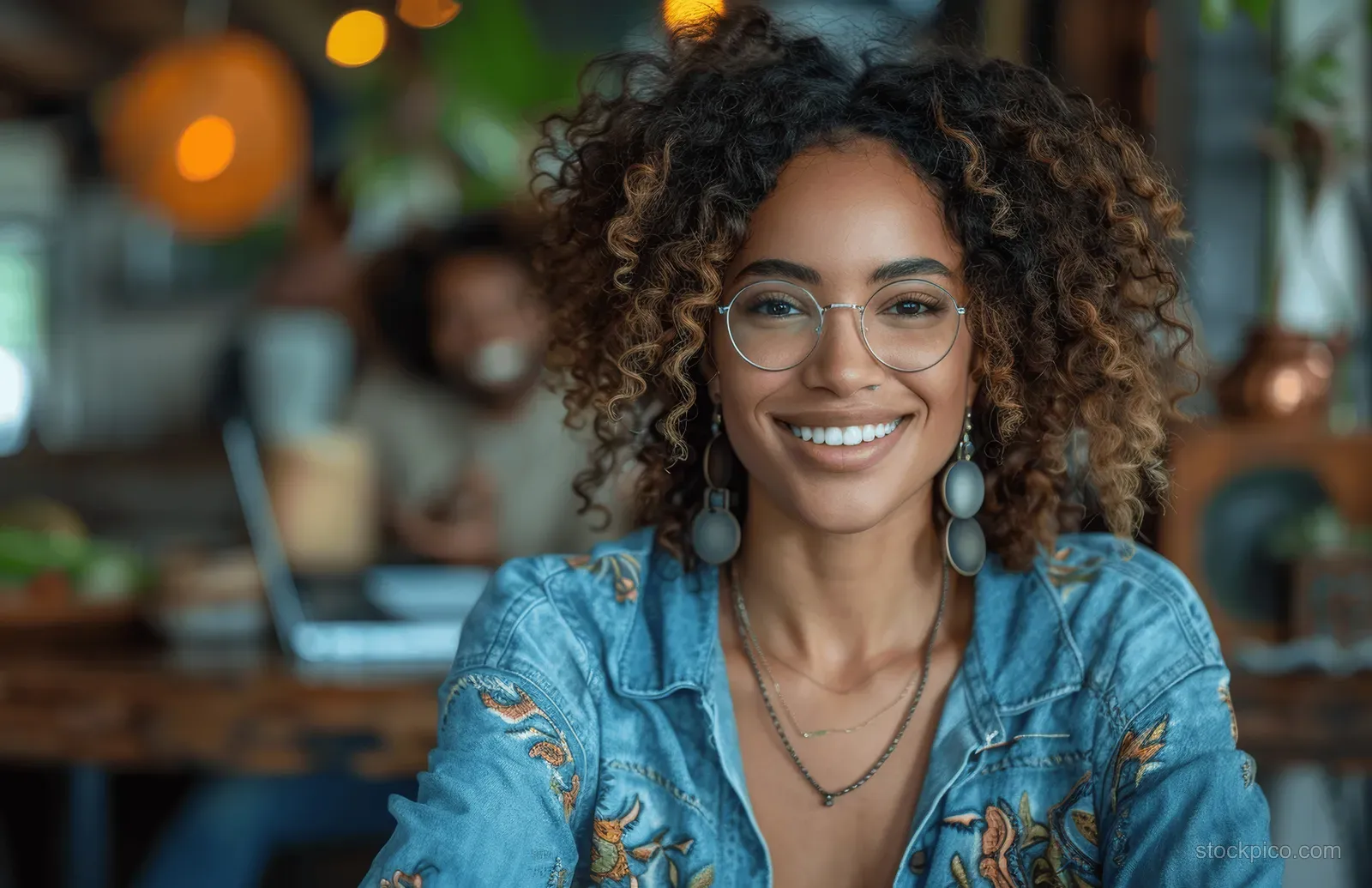 Smiling woman with curly hair and glasses works on a laptop at an office conference table with blurred coworkers behind.