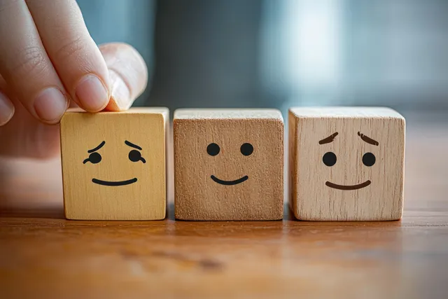 A hand holds a wooden block with a smiling face while two other emotion blocks sit on a table.