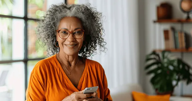 Happy senior Black woman with curly hair and glasses using smartphone in bright living room
