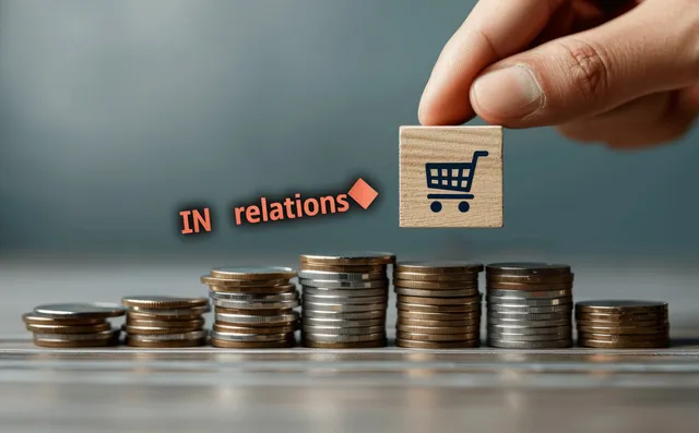 Hand holding a wooden block above stacked coins with a shopping cart icon suggesting rising inflation costs.