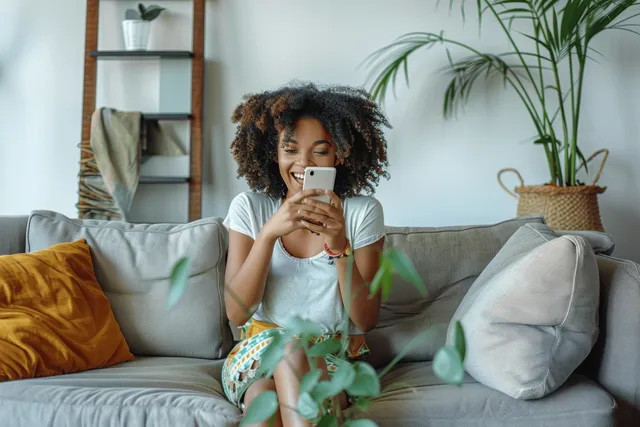 Excited Black woman smiling while holding smartphone and celebrating on gray couch at home