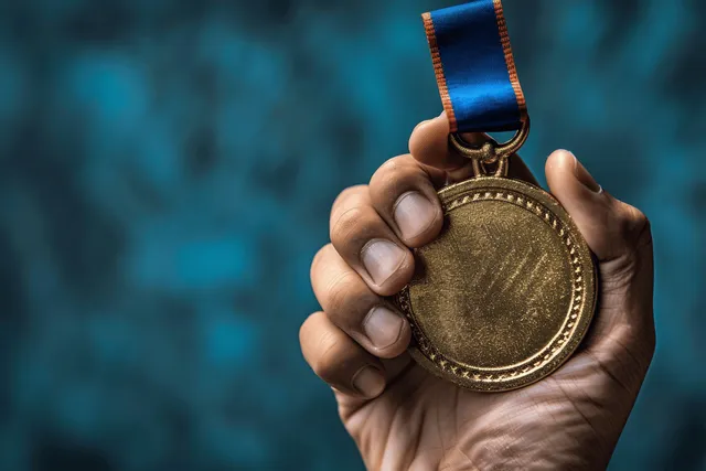 A hand holds a gold medal with a blue ribbon against a blurred blue background.