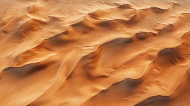 Aerial view of rolling sand dunes with warm orange color and soft shadows across a sunlit desert.