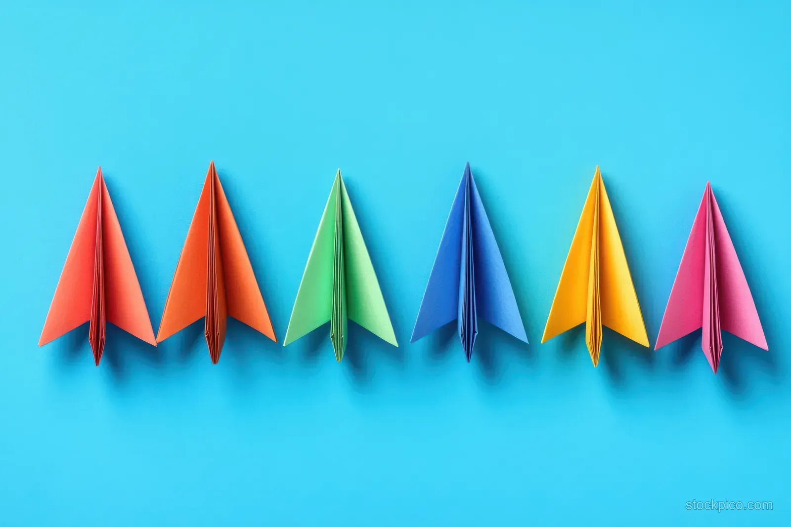 Multiple colorful paper airplanes line up on a bright blue background, suggesting competition and forward flight.