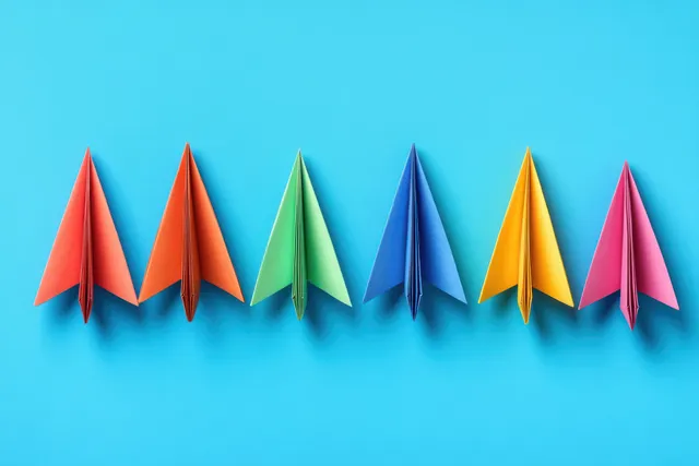 Multiple colorful paper airplanes line up on a bright blue background, suggesting competition and forward flight.