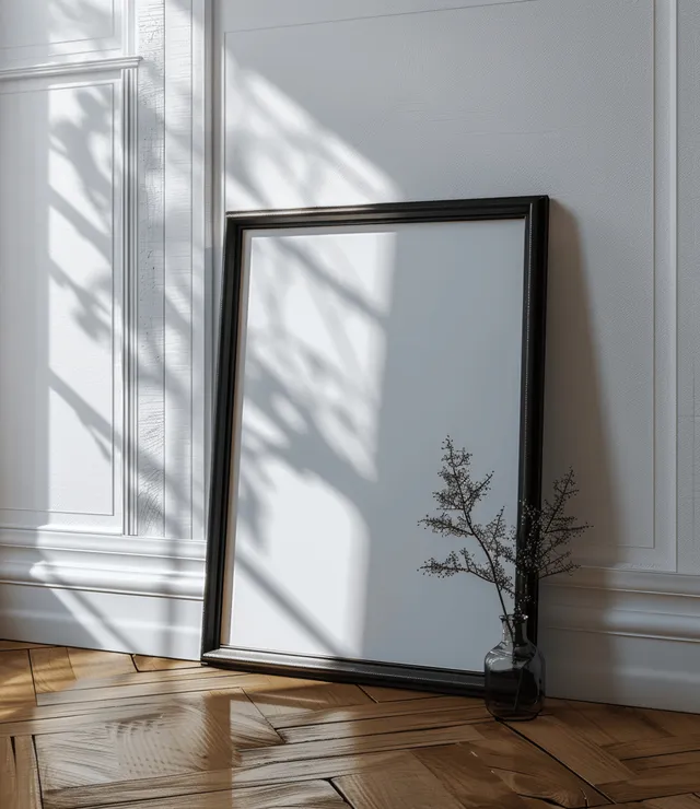 Empty black photo frame leaning against a white wall on parquet flooring with window light reflections.