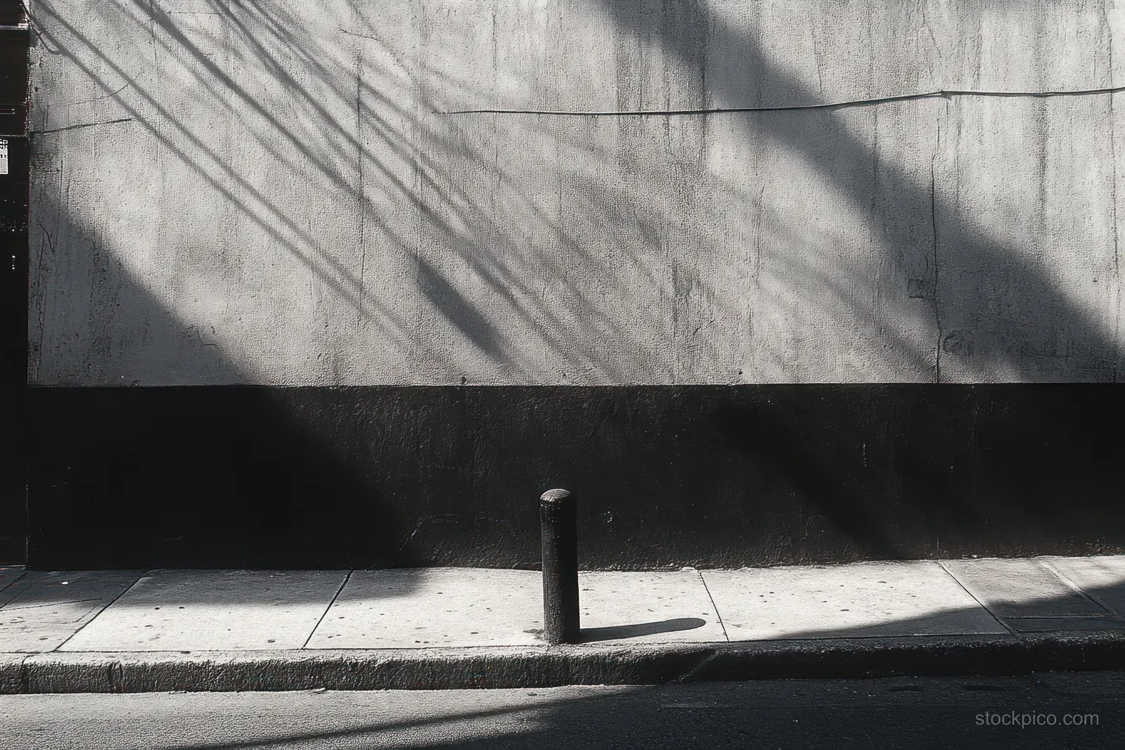 Empty monochrome sidewalk beside a textured concrete wall with a pole and intersecting shadows cast by sunlight.