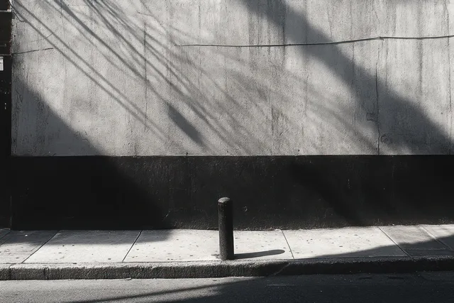 Empty monochrome sidewalk beside a textured concrete wall with a pole and intersecting shadows cast by sunlight.