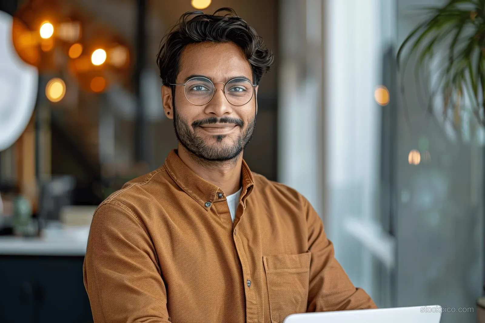 Smiling young man in glasses typing on a laptop at a desk in a modern office with warm bokeh lights.