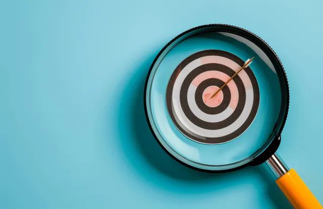 Magnifying glass reveals a dartboard bullseye with an arrow on a blue background.