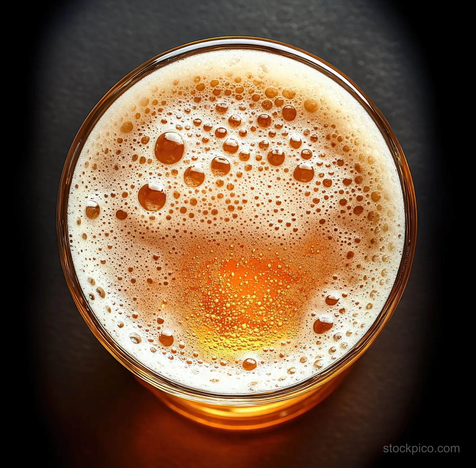 Top view of amber beer with frothy white bubbles in a glass on a dark surface.