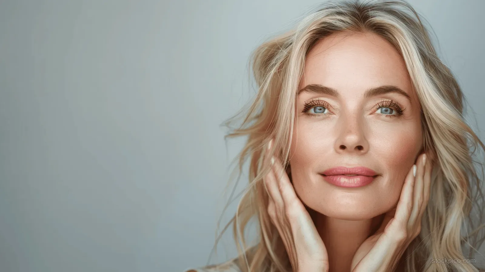 Blonde woman in a close-up portrait gently touching her cheeks with a calm blue-eyed expression.