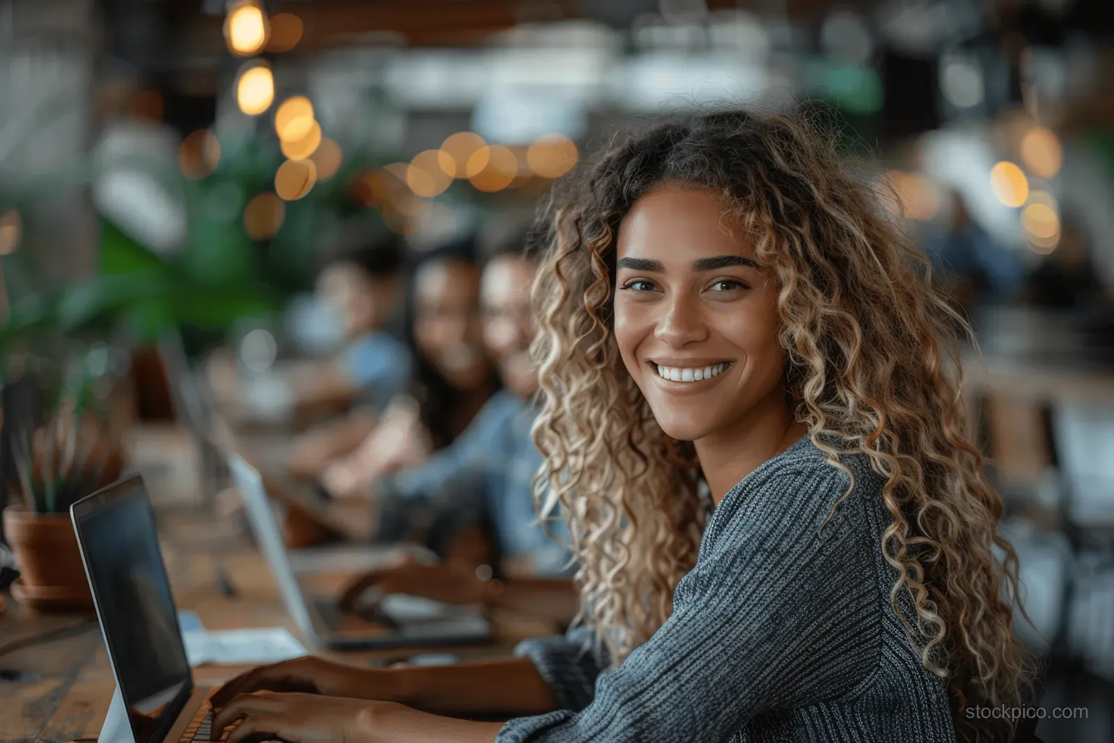 Smiling businesswoman works on a laptop in a shared open office with blurred desks and coworkers behind.