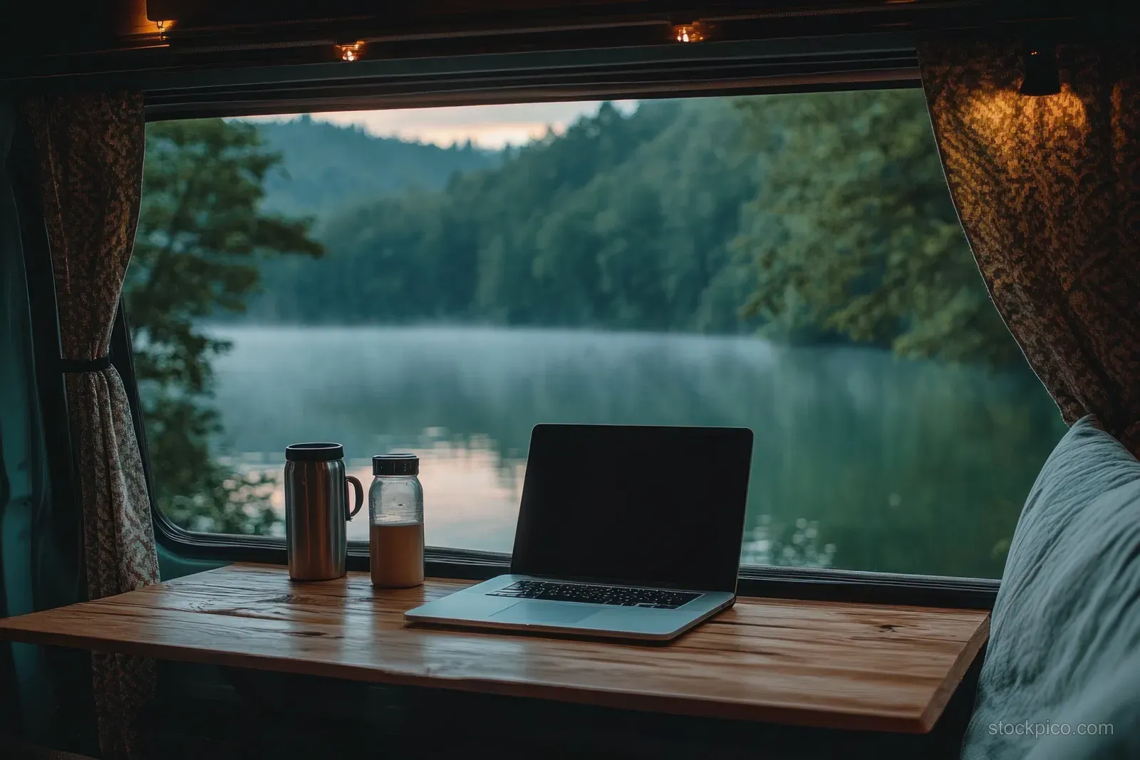 Laptop on a wooden table inside a camper van beside a travel mug and bottle, framed by curtains overlooking a misty lake and forest.