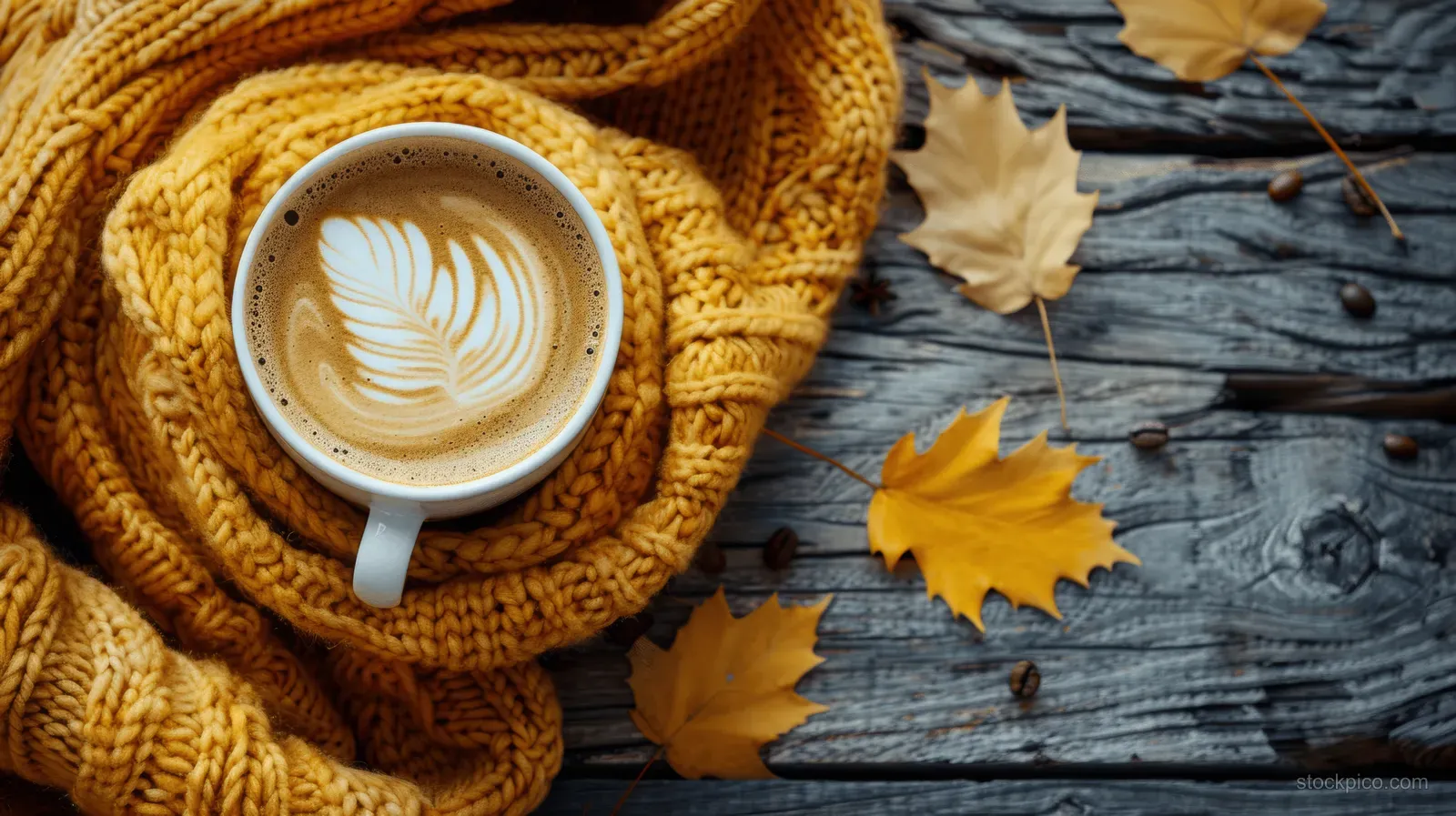 A latte mug wrapped in a knitted scarf on a rustic wooden table with scattered autumn leaves.