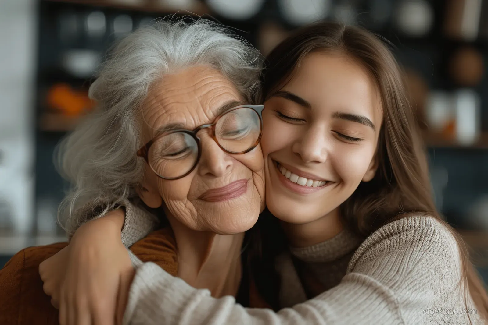A young daughter hugs her smiling senior mother at home, their faces close in a warm affectionate embrace.