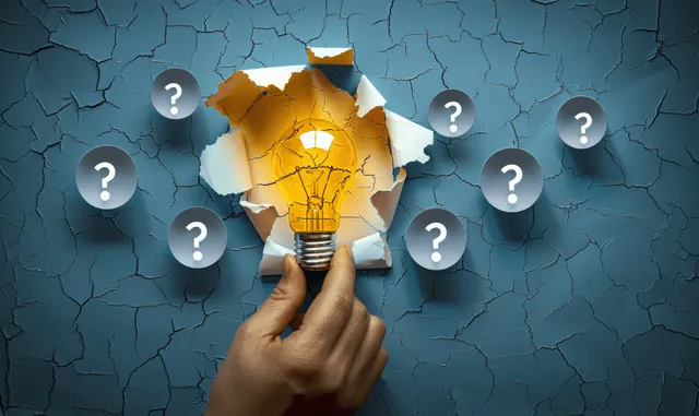 A hand tears torn paper to reveal a glowing light bulb with question marks on a cracked blue wall.