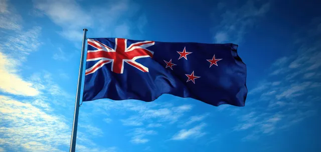 A New Zealand flag waves on a pole against a bright blue sky with scattered clouds.