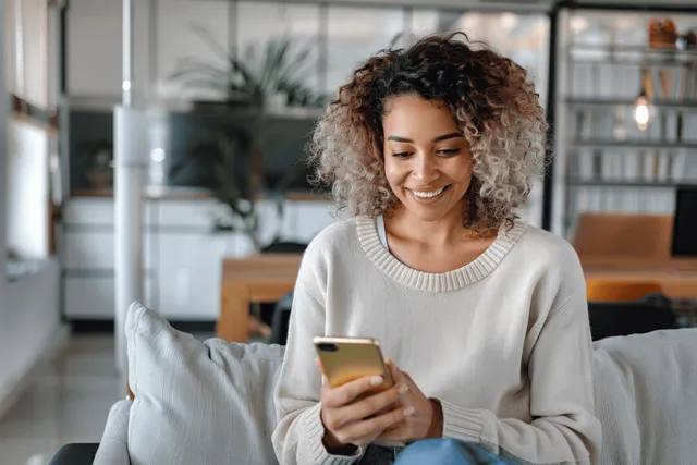 Happy mixed-race woman on sofa texting on a yellow smartphone in a bright minimal living room