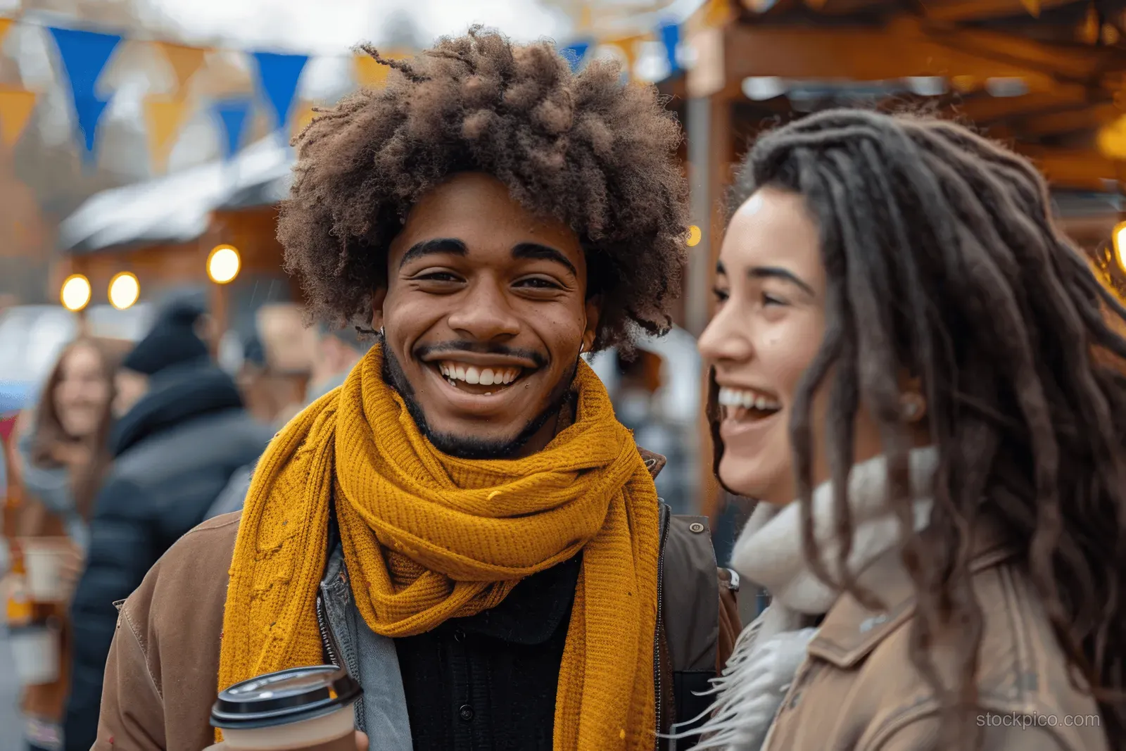 Two young friends laugh while holding coffee cups at an autumn street market with flags and stalls behind them.