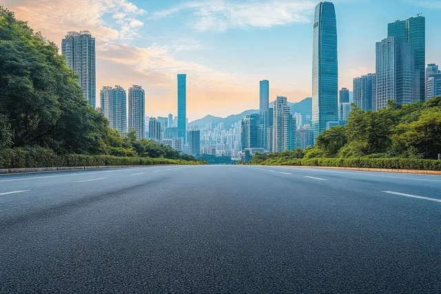 An empty asphalt road runs between green trees toward tall modern skyscrapers under a partly cloudy sunset sky.