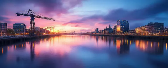 Calm river reflects a purple and orange sunset above a modern skyline with cranes and contemporary buildings.