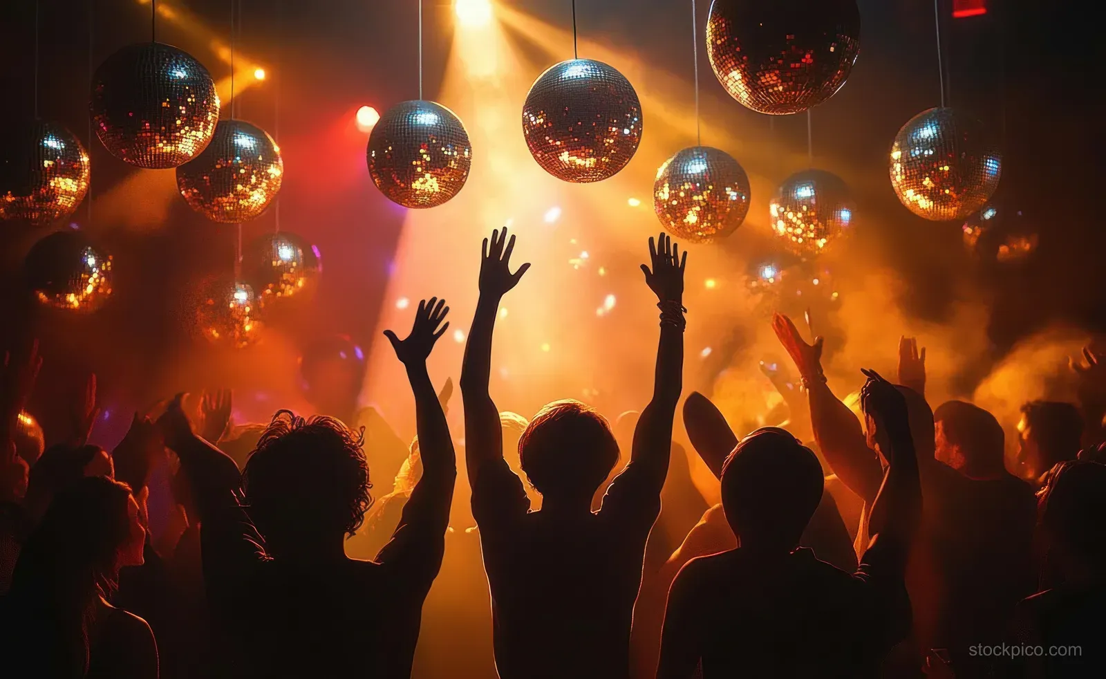 A crowd dances with raised hands under hanging disco balls in a warm orange-lit nightclub.