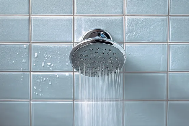 A chrome showerhead sprays water onto a light blue tiled wall with droplets visible.