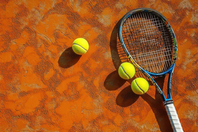 Tennis racket and three bright yellow balls on orange clay court casting distinct shadows