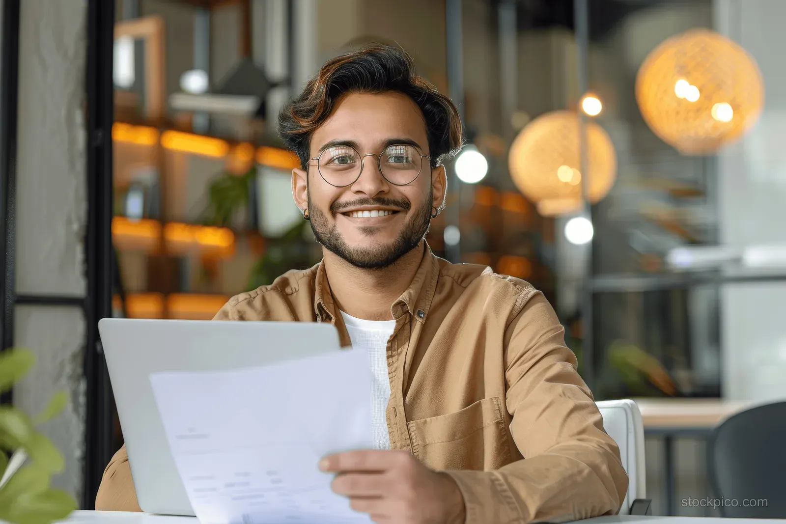 Smiling young man holding documents at a desk with a laptop in a modern office with warm lights.
