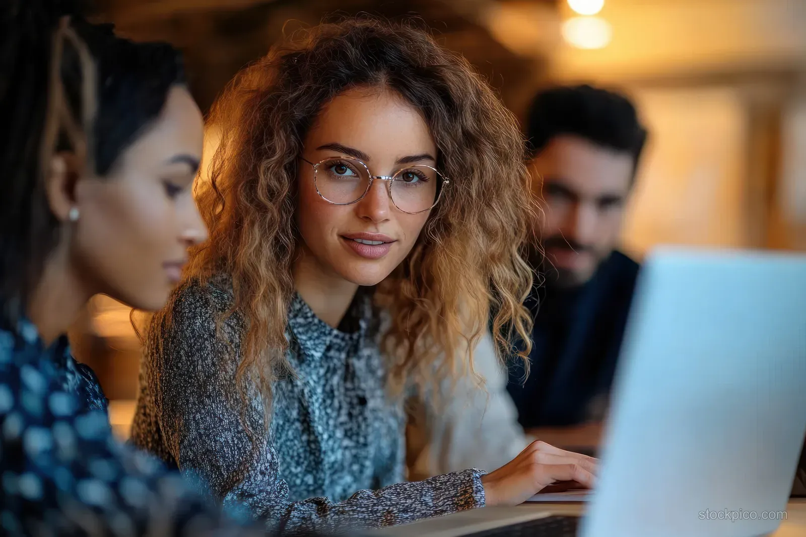 A woman with glasses smiles while typing on a laptop as coworkers collaborate around desks indoors.