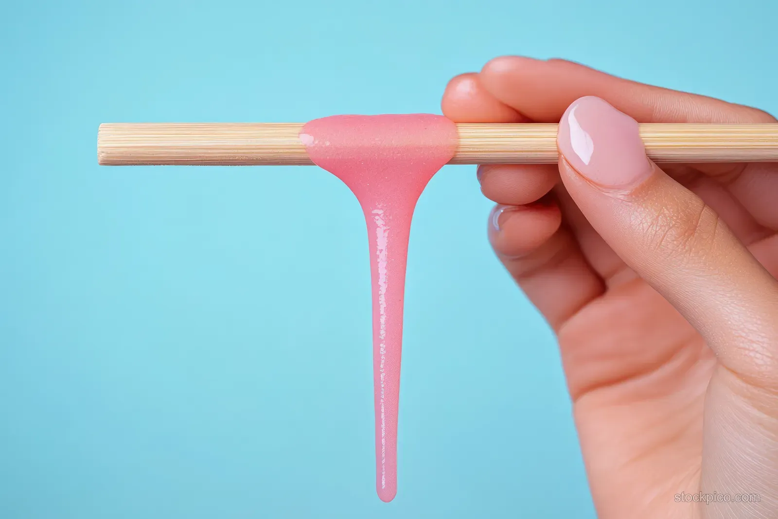 A hand holds a depilation wax stick with pink wax dripping against a blue background.