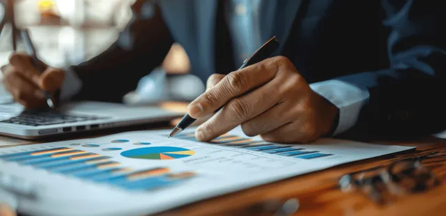 Businessman’s hand writes on printed financial charts on an office desk beside a laptop.