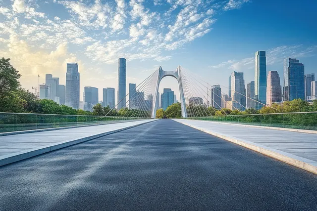 Suspension bridge road curves toward a city skyline with trees along the sides under a blue sky.