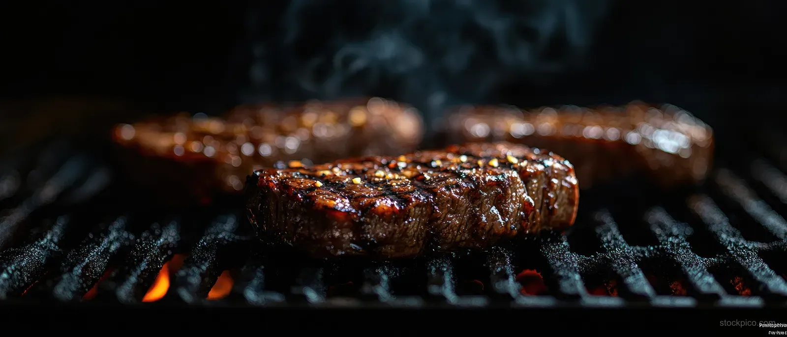 Beef steak sizzling on a grill with visible char marks and smoke, herbs and hands at the side.