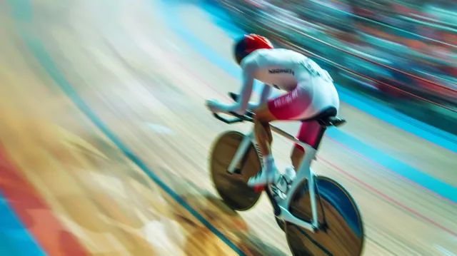 A male track cyclist speeds along a blue velodrome turn, body leaning forward with pronounced motion blur.