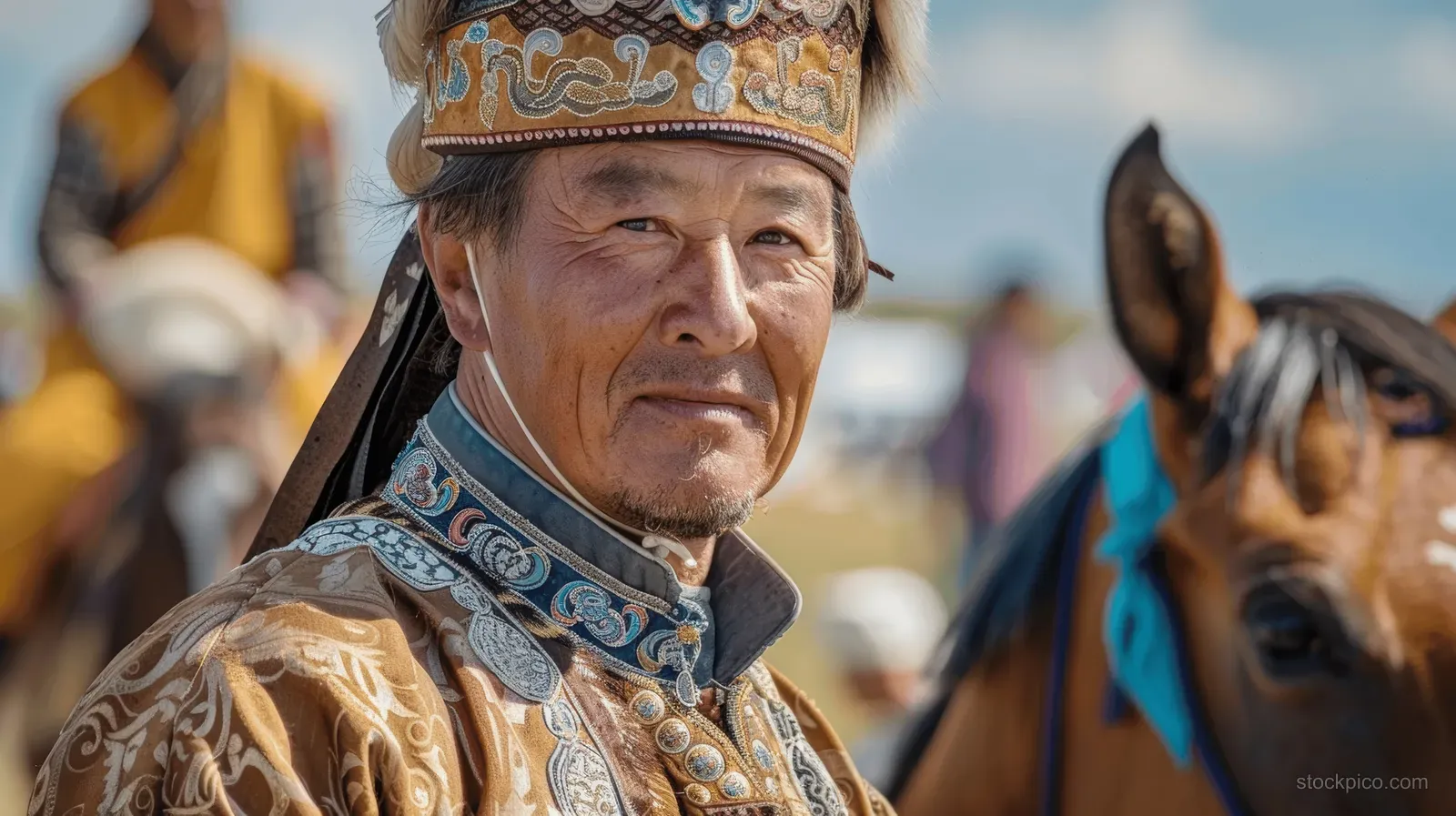 A Mongolian rider in an ornate hat stands beside a dappled horse at a Naadam festival in daylight.