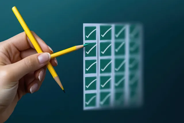 A hand holds a yellow pencil beside a checklist paper with green checkmarks on a blue background.