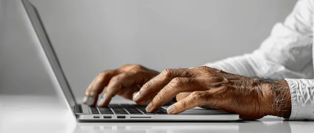 Closeup of hands typing on a laptop while using a smartphone on a modern office desk with a clean white background.