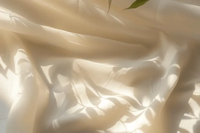 Beige fabric close up with soft folds and sunlit leaf-shaped shadows on the cloth.