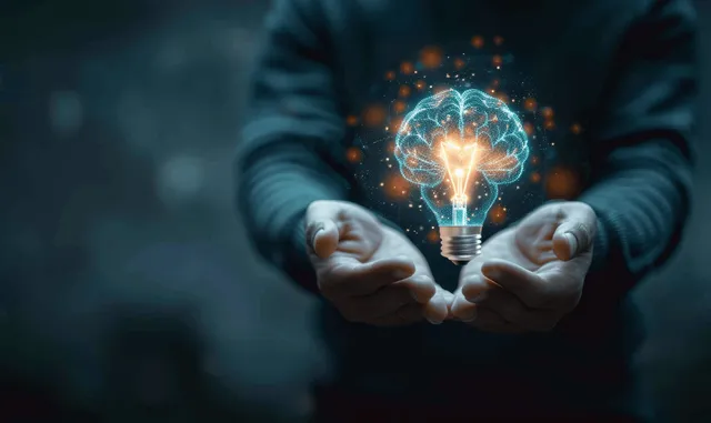 A businessman holds a glowing light bulb projecting a brain icon hologram in a dark blurred background.