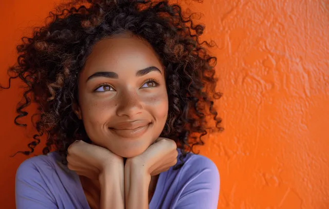 Woman with curly hair covers her face with hands and smiles against a textured orange wall.