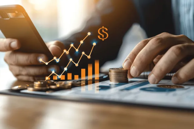 A businessman uses a smartphone to review ROI and growth while coins and a chart lie on a desk.