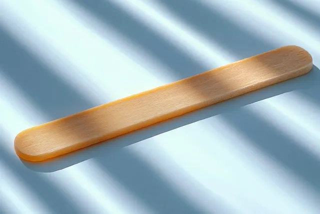 A wooden tongue depressor rests on a light blue surface with diagonal shadows and clear wood texture.