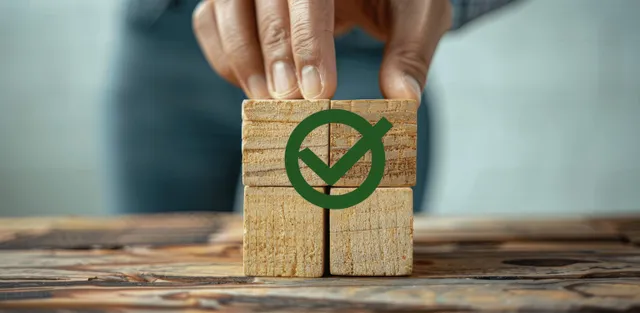 A hand places a wooden cube with a green checkmark ISO-style symbol on a table.