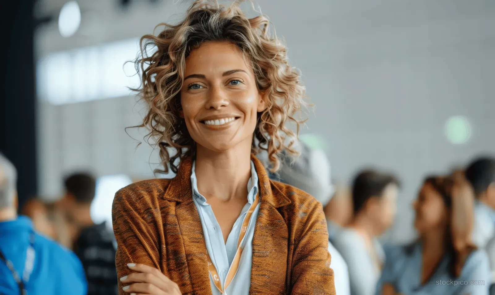 A smiling curly-haired woman stands indoors wearing a casual business outfit.