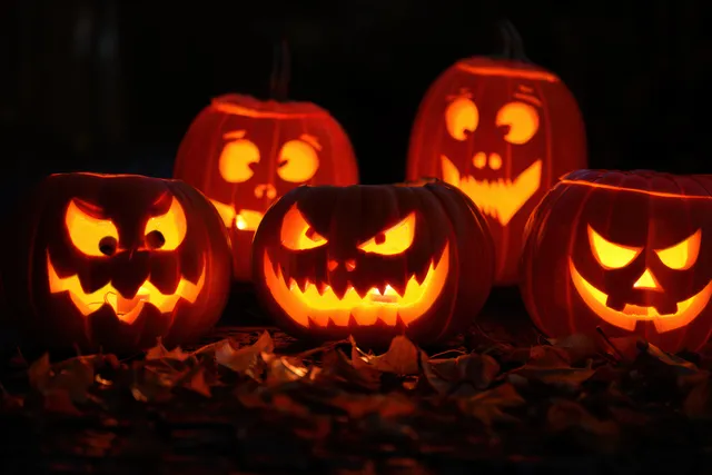 Five glowing carved jack-o-lantern pumpkins with spooky faces sit on scattered autumn leaves in the dark.