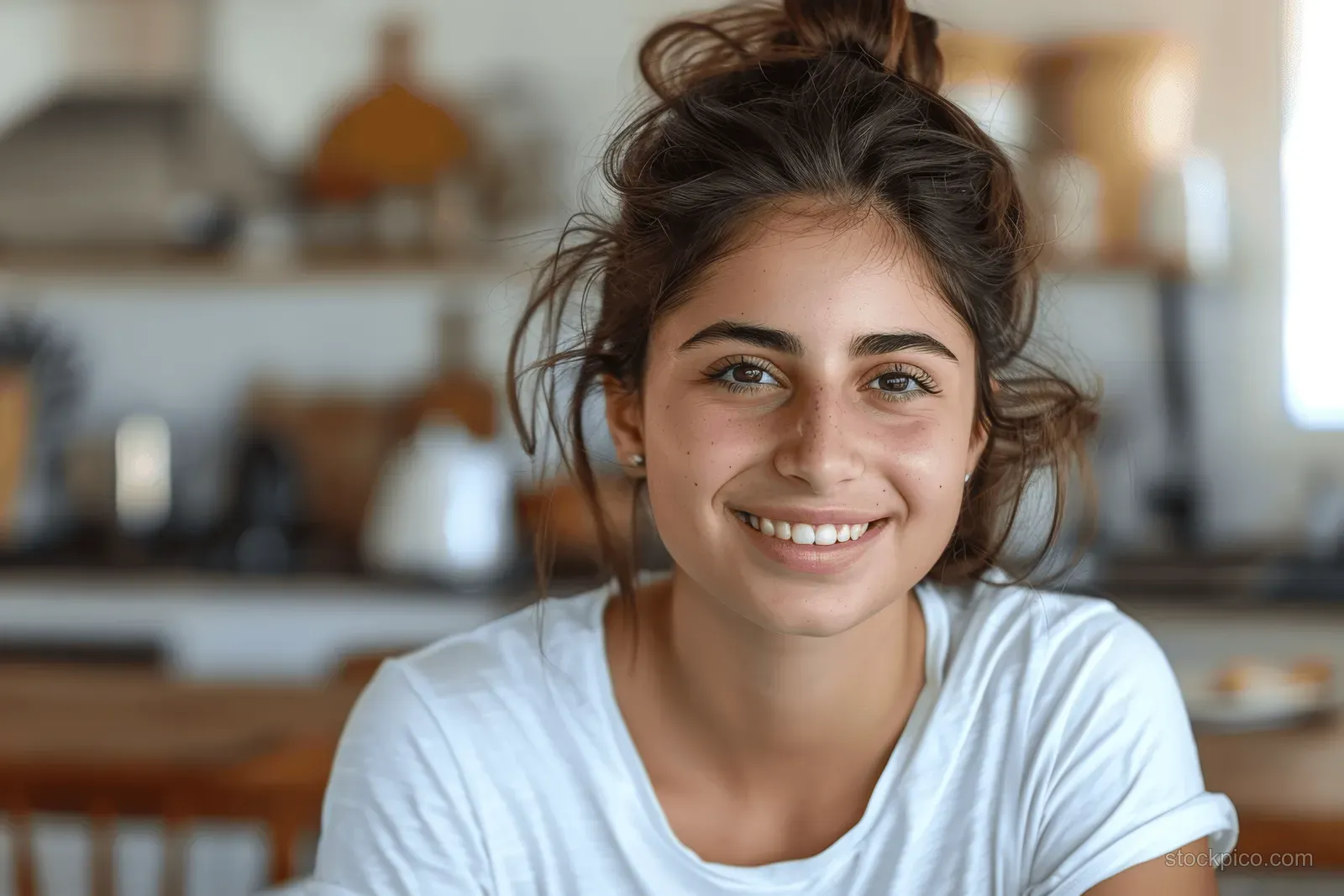 Smiling young woman in a white T-shirt stands indoors in a cozy kitchen with warm natural light.