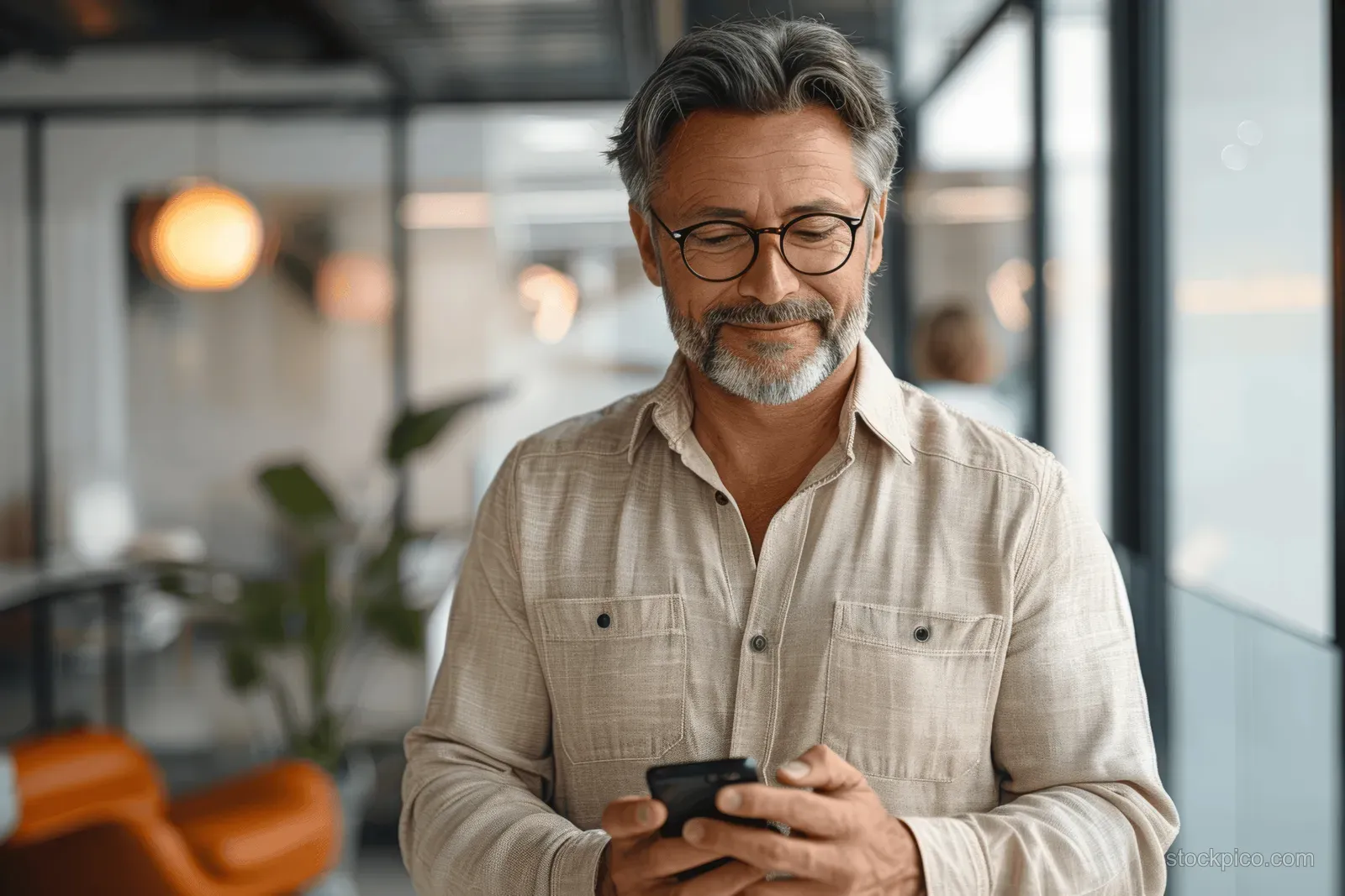 A smiling mature businessman in an office holds a smartphone and taps an app on the screen.