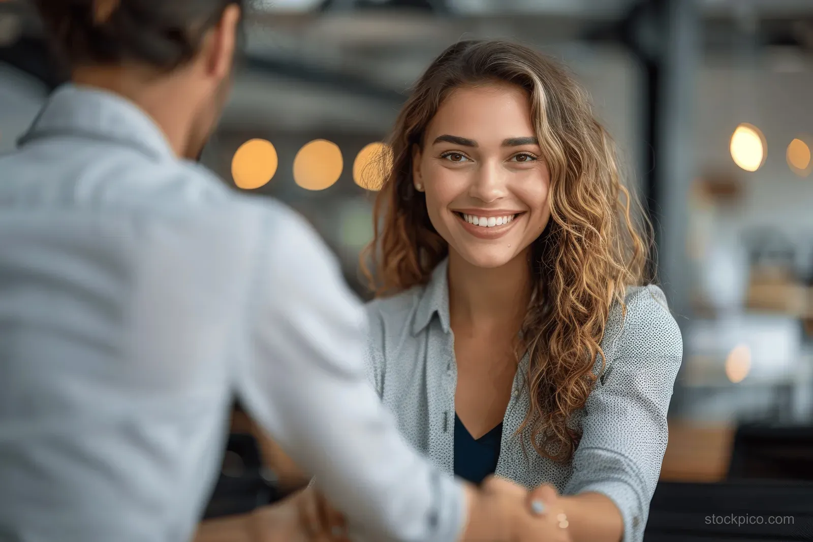 Smiling businesswoman shakes hands with a coworker across an office meeting table with warm bokeh lights behind.
